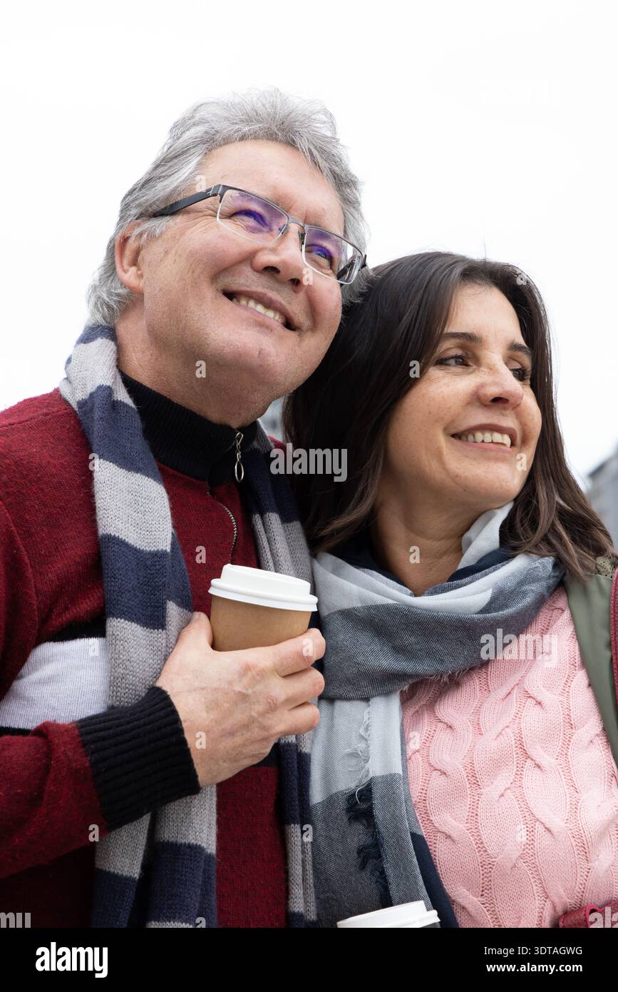 Couple d'âge moyen debout dehors sous le ciel nuageux, tenant des tasses à emporter et portant des foulards Banque D'Images