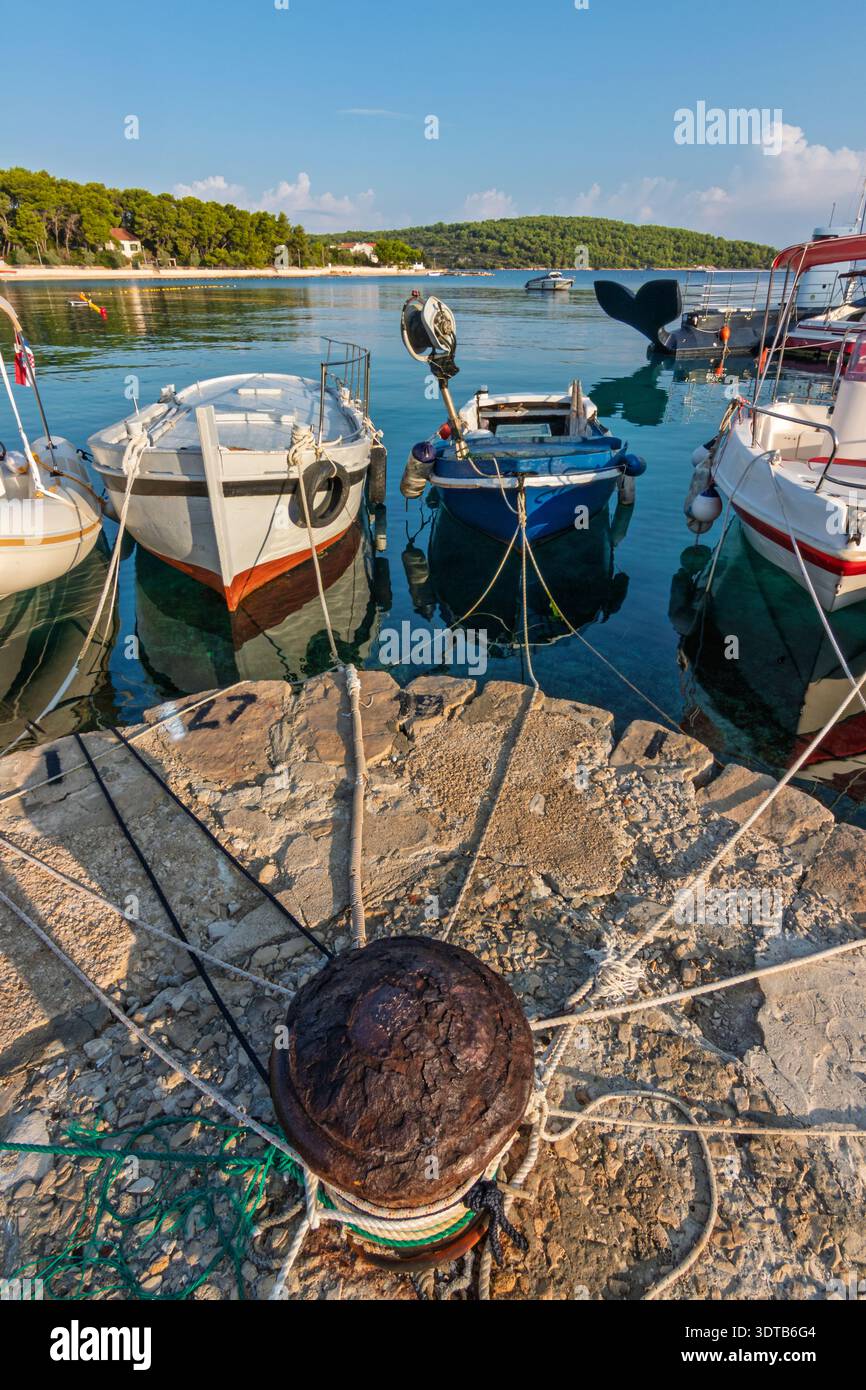 Petits bateaux de pêche et embarcations de plaisance amarrés à un quai rocheux avec des cordes et des crampons, reflétés dans une eau turquoise claire sous un ciel ensoleillé à côté de pins Banque D'Images