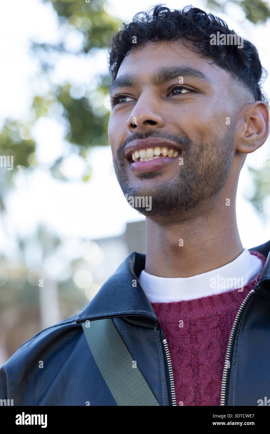 Homme indien souriant, portant une veste en cuir noir et un pull marron avec sangle verte dans le parc Banque D'Images