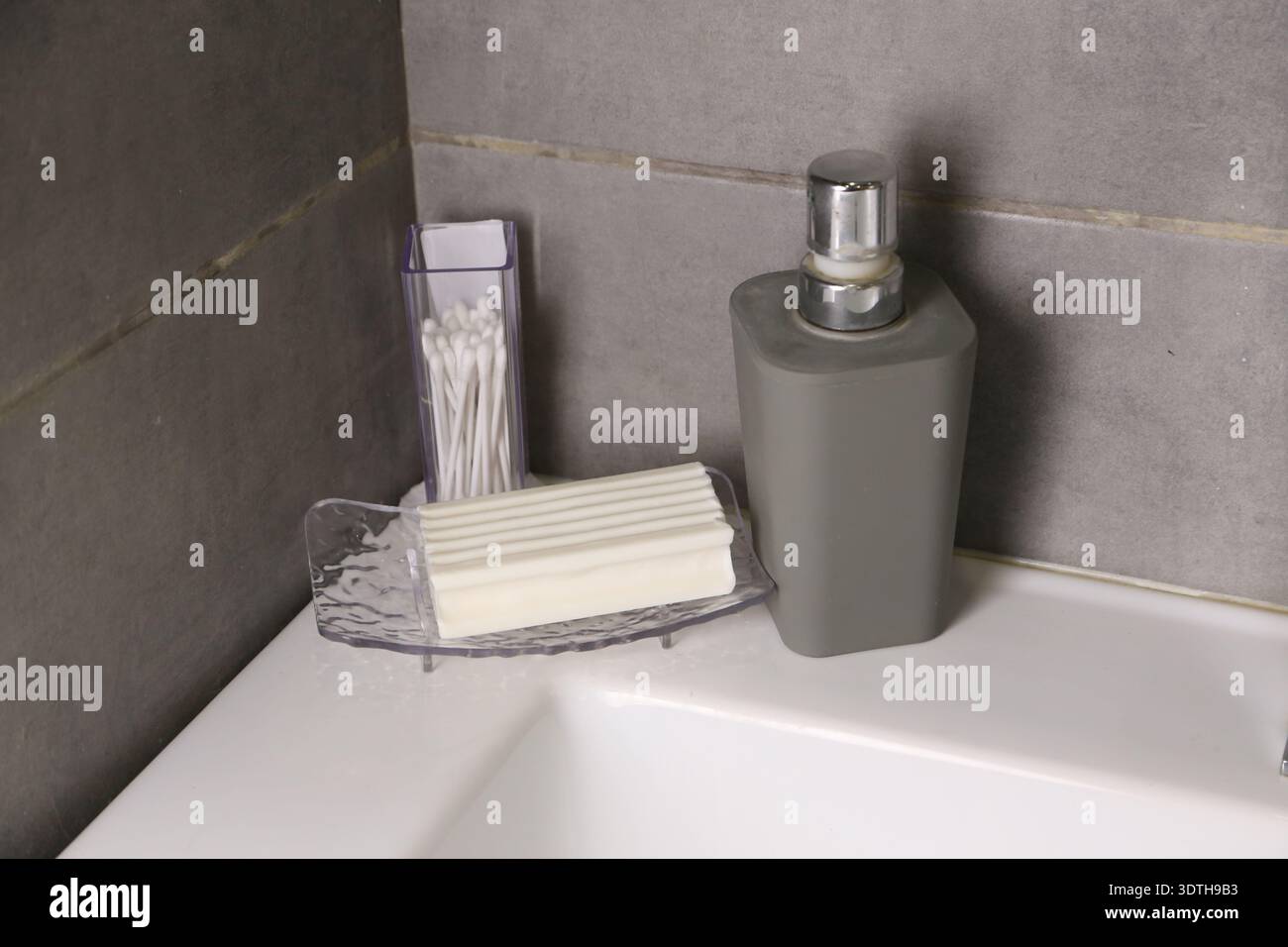 Un coin d'une salle de bain montre un porte-savon avec une barre de savon, un récipient avec du savon liquide et un support rempli de cotons-tiges. La lumière remplit l'espace pendant la journée. Banque D'Images
