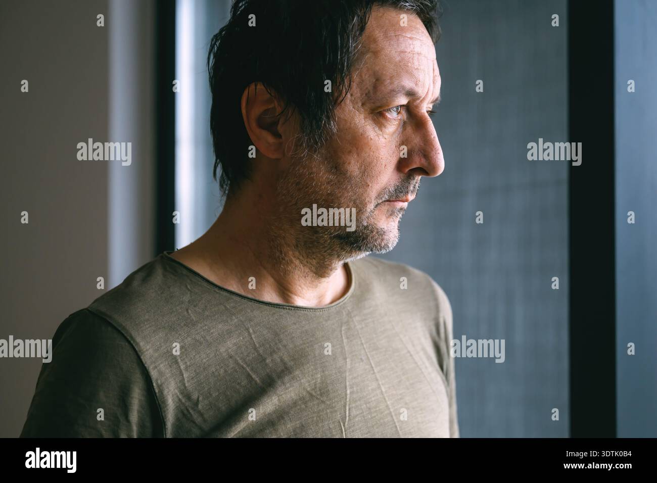 Homme adulte mûr avec les cheveux unkempt et chaume facial regardant par la fenêtre de son appartement, expression triste et déçue, indoo réfléchissant calme Banque D'Images