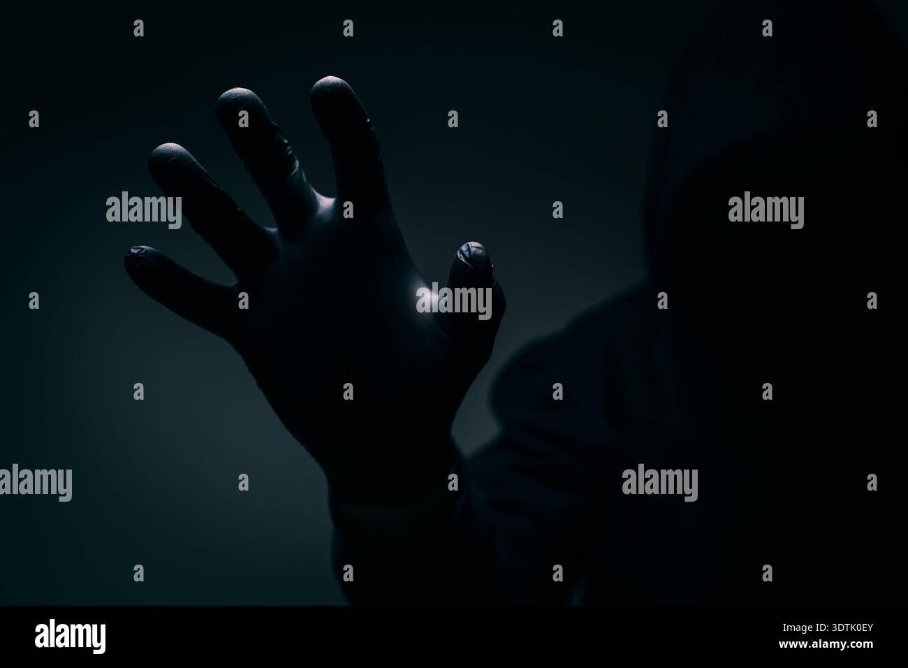 Silhouette à capuche sombre étendant une main gantée vers le spectateur, éclairage dramatique véhiculant le danger, la peur, la tension et l'anonymat. Mise au point sélective. Banque D'Images