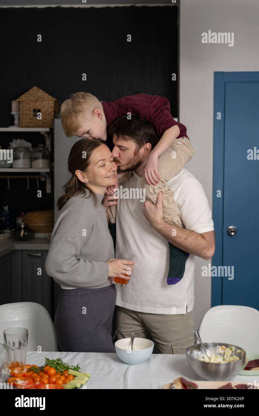 Une famille aimante partage un moment tendre dans la cuisine alors que le père embrasse la mère tout en tenant son enfant sur ses épaules, entouré de frais Banque D'Images