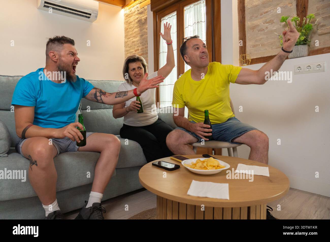 Trois amis réagissent passionnément tout en regardant un match de sport à la maison, acclamant et se disputant avec des bières et des chips sur la table dans un salon animé Banque D'Images