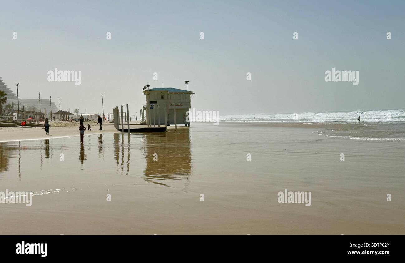 Tour de sauveteur reflétée dans le sable humide sur une plage brumeuse. Les gens marchant le long du rivage avec des vagues douces de l'océan dans une scène côtière calme et atmosphérique - Image de stock capturée avec un smartphone