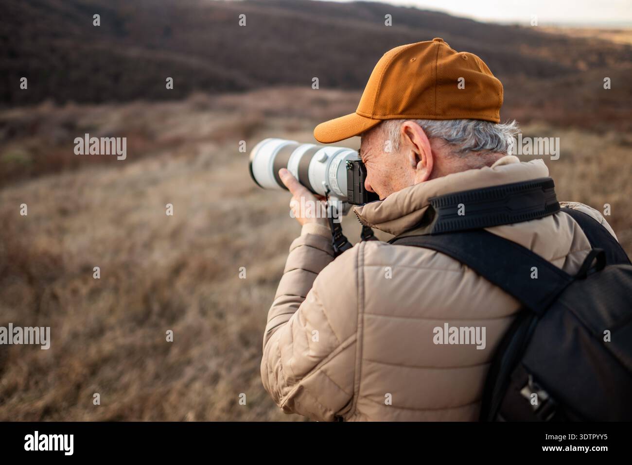 Un homme portant une casquette, une veste rembourrée et un sac à dos debout dans la montagne, se concentrant à travers son objectif de caméra pour photographier quelque chose. Banque D'Images