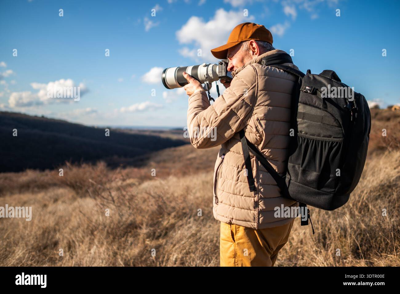 Un homme portant une casquette, une veste rembourrée et un sac à dos debout dans la montagne, se concentrant à travers son objectif de caméra pour photographier des paysages ou la faune. Banque D'Images