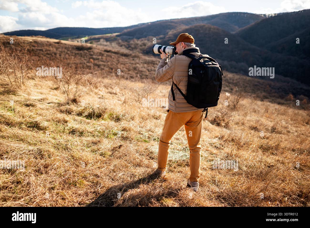 Un homme portant une casquette, une veste rembourrée et un sac à dos debout dans la montagne, se concentrant à travers son objectif de caméra pour photographier des paysages ou la faune. Banque D'Images