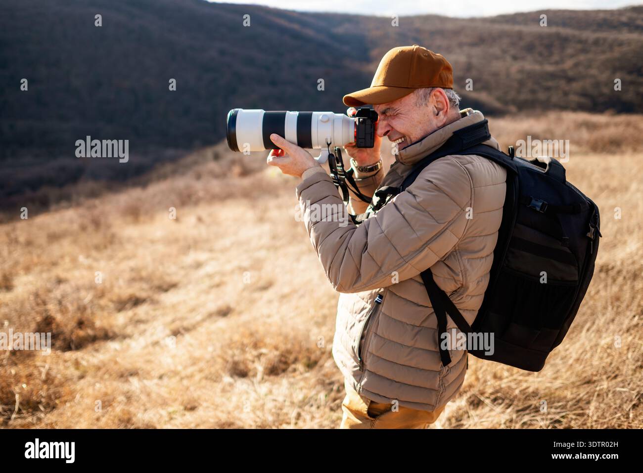 Un homme portant une casquette, une veste rembourrée et un sac à dos debout dans la montagne, se concentrant à travers son objectif de caméra pour photographier des paysages ou la faune. Banque D'Images