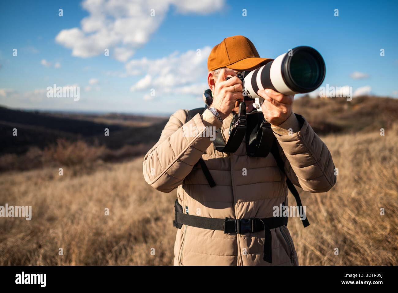 Vue rapprochée d'un photographe animalier visant un grand téléobjectif à travers un paysage de campagne ouvert sous un ciel bleu vif. Banque D'Images