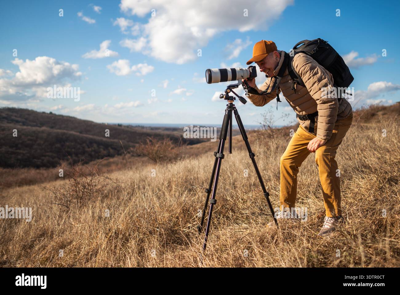 Photographe animalier senior utilisant un trépied et visant un grand téléobjectif à travers un paysage de campagne ouvert par jour ensoleillé. Banque D'Images