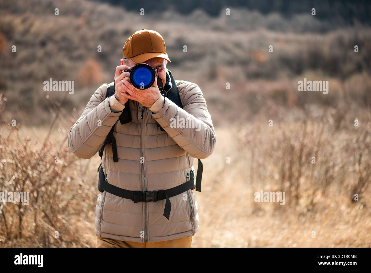 Un homme portant une casquette, une veste rembourrée et un sac à dos debout dans une prairie d'herbe sèche, se concentrant à travers son objectif de caméra pour photographier quelque chose. Banque D'Images