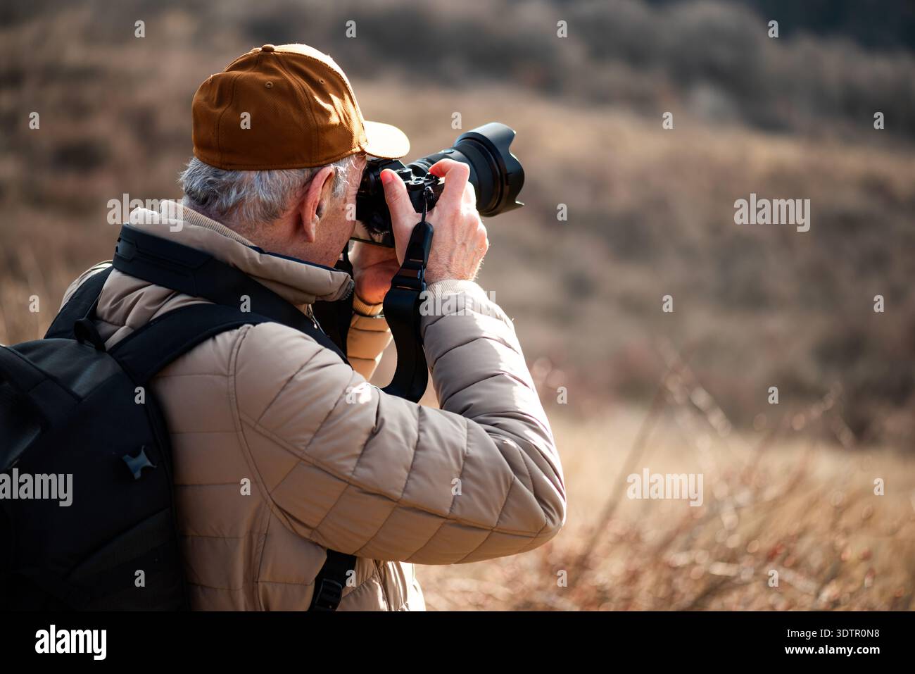 Un homme portant une casquette, une veste rembourrée et un sac à dos debout dans une prairie d'herbe sèche, se concentrant à travers son objectif de caméra pour photographier quelque chose. Banque D'Images