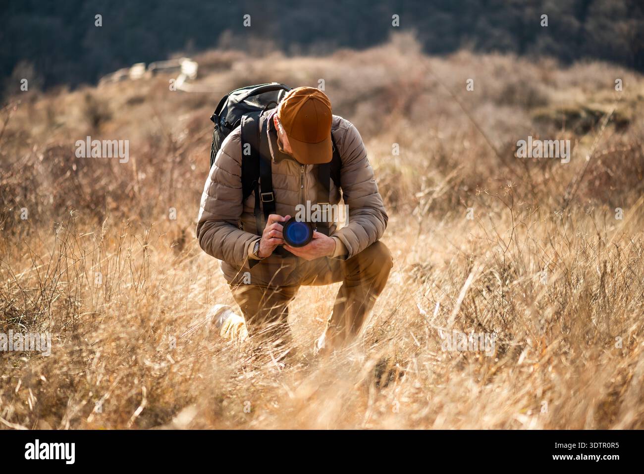 Un photographe de la nature s'agenouille dans un champ d'herbe sèche, ajustant soigneusement les paramètres de son appareil photo tout en capturant des détails rapprochés du paysage en guerre Banque D'Images