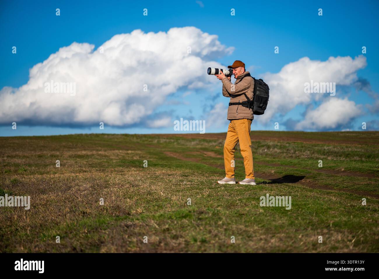 Un homme portant une casquette marron, une veste rembourrée et un sac à dos prend des photos de la faune ou du paysage à l'extérieur, entouré de champs roulants et de cl blanc spectaculaire Banque D'Images