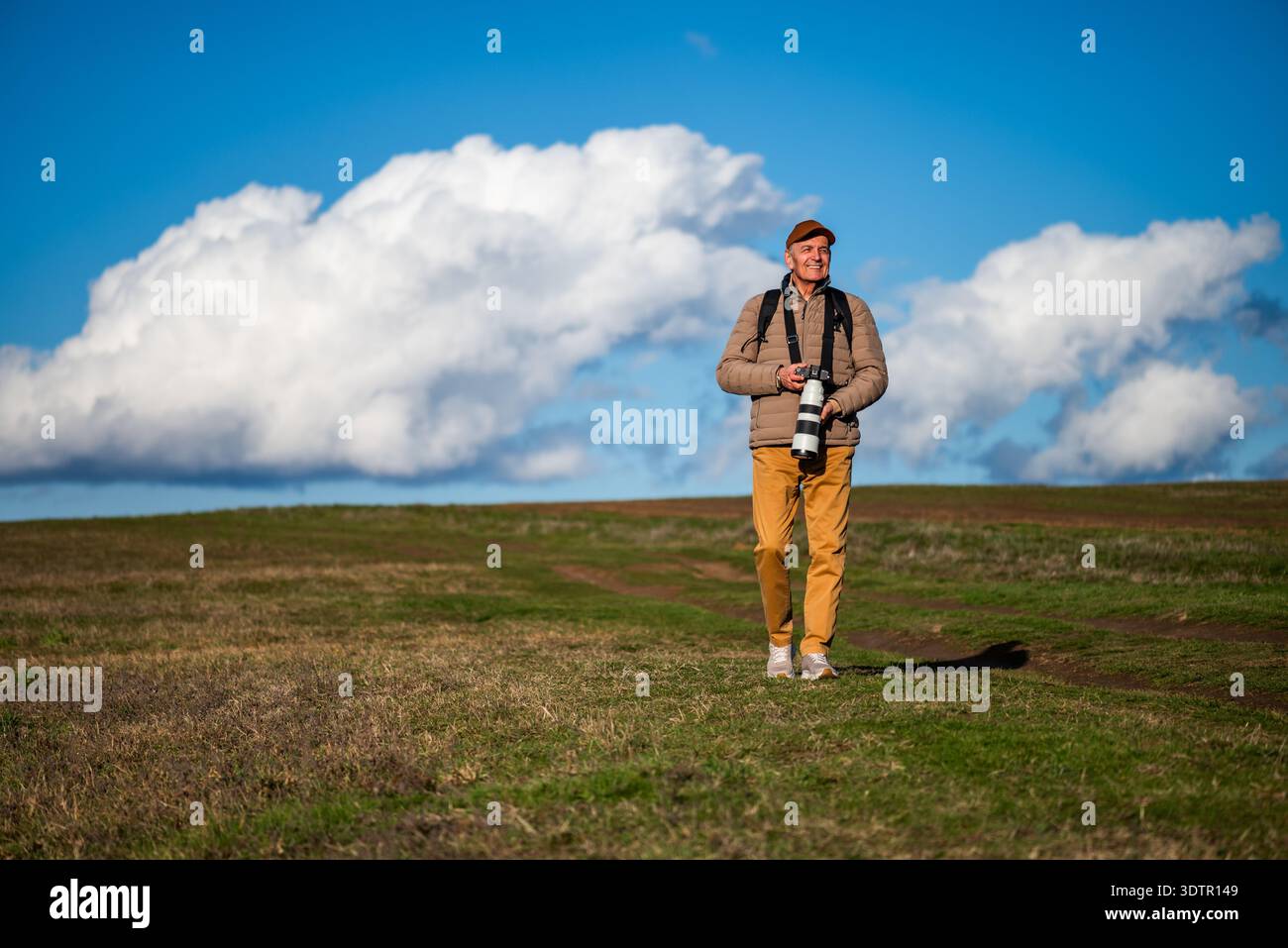 Un photographe masculin mature se tient dans un champ herbeux ouvert sous un ciel bleu vif, capturant des sujets éloignés avec un appareil photo professionnel équipé de Banque D'Images