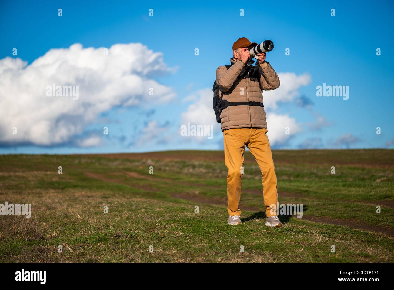 Un homme portant une casquette marron, une veste rembourrée et un sac à dos prend des photos de la faune ou du paysage à l'extérieur, entouré de champs roulants et de cl blanc spectaculaire Banque D'Images