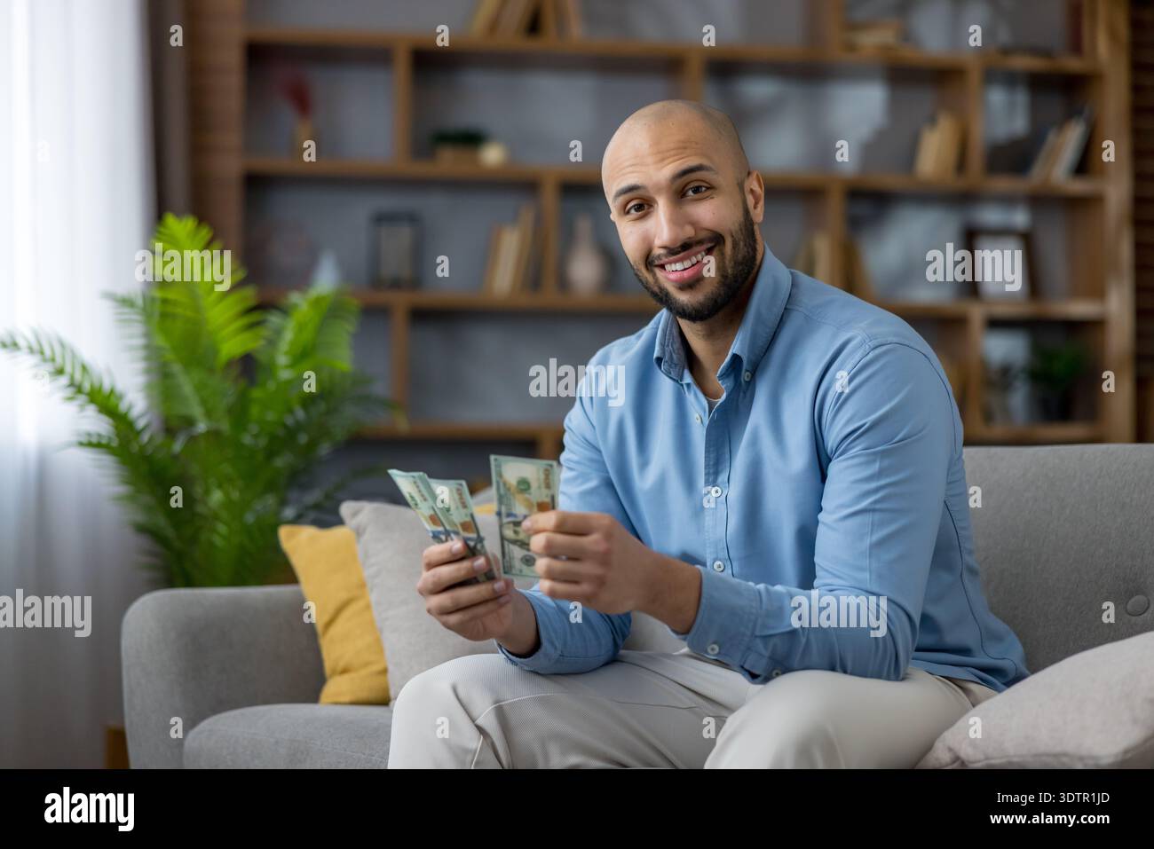 Jeune homme joyeux souriant, tenant des piles de billets de banque en dollars, gérant avec succès son budget et comptant l'argent tout en étant assis sur un canapé confortable dans un salon moderne Banque D'Images
