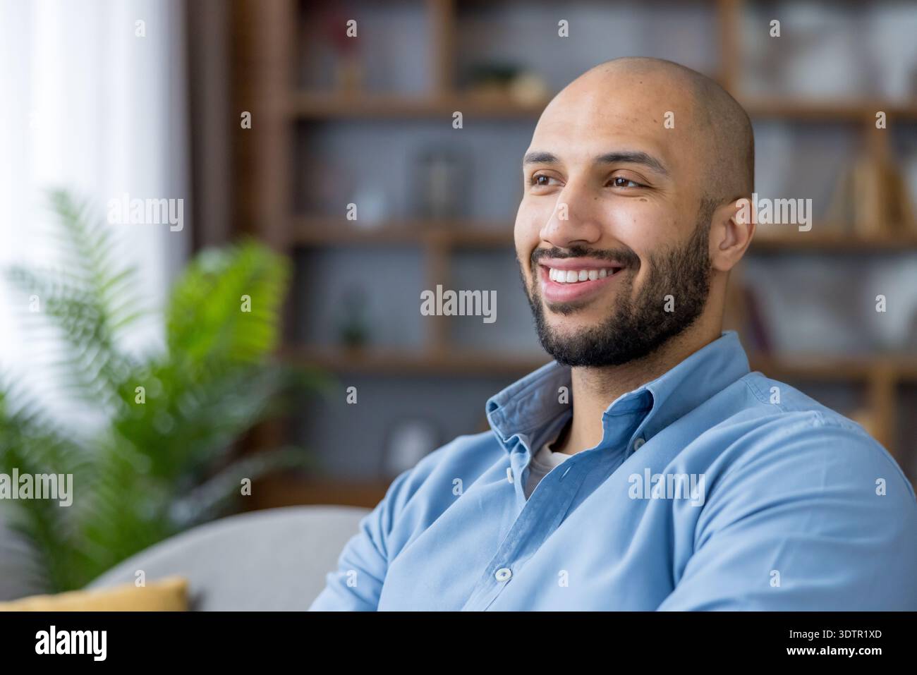 Homme chauve et barbu souriant de profil, assis détendu dans un salon lumineux, regardant loin avec joie et contentement, incarnant la confiance détendue et l'équilibre travail-vie privée Banque D'Images