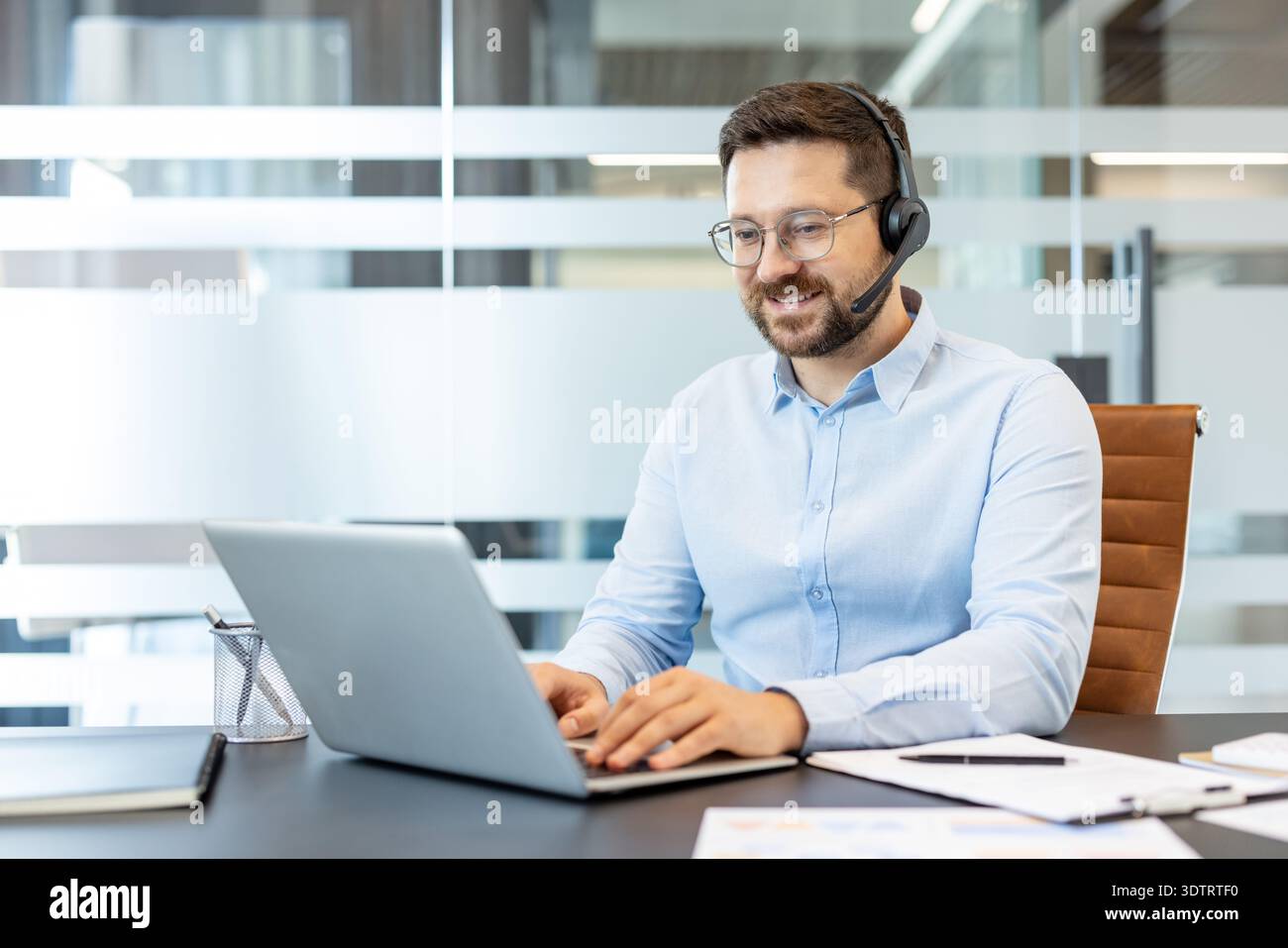 Jeune homme portant un casque avec un microphone et une chemise bleu clair assis à un bureau moderne, tapant sur un ordinateur portable, fournissant un service à la clientèle et une assistance Banque D'Images