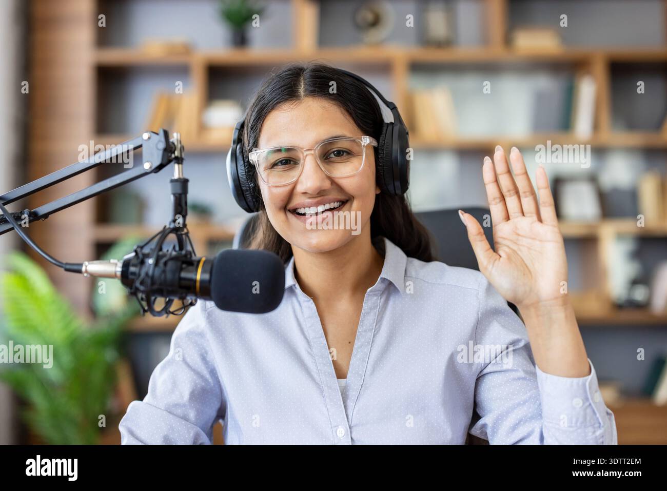Femme heureuse portant des écouteurs et des lunettes, enregistrant un podcast avec un microphone professionnel, agitant la main tout en diffusant une session audio en direct depuis un home studio Banque D'Images