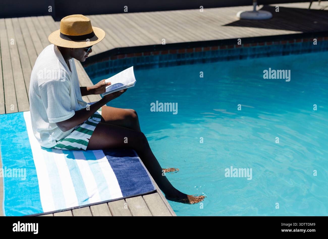Livre de poche ouvert reposant sur une serviette bleue et blanche au bord de la piscine avec chapeau de paille et lunettes de soleil Banque D'Images