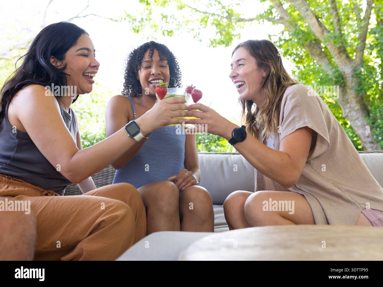 Diverses femmes amies riant et grillant avec des boissons à la fraise sur le canapé extérieur, portant des montres Banque D'Images