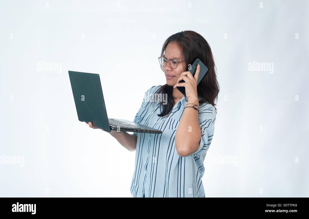 Femme indonésienne portant des lunettes et une chemise rayée bleue, semblant stressée et concentrée tout en tenant un ordinateur portable et en parlant sur son smartphone à la sa Banque D'Images