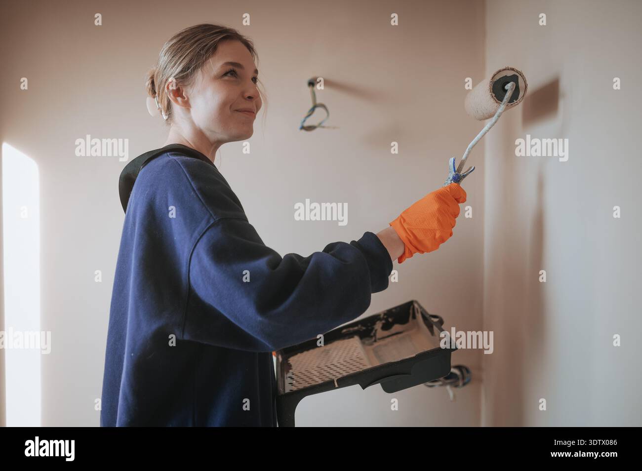 Une jeune femme peint en toute confiance le mur d'une pièce à l'aide d'une brosse rotative. Portant des gants de protection et tenant un bac à peinture, elle ajoute un nouveau manteau à Banque D'Images