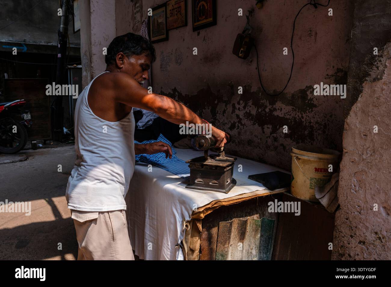 Un homme dans un gilet blanc utilise un fer lourd traditionnel pour presser une chemise à damier bleue sur une table simple dans une ruelle étroite dans le vieux Delhi. Banque D'Images