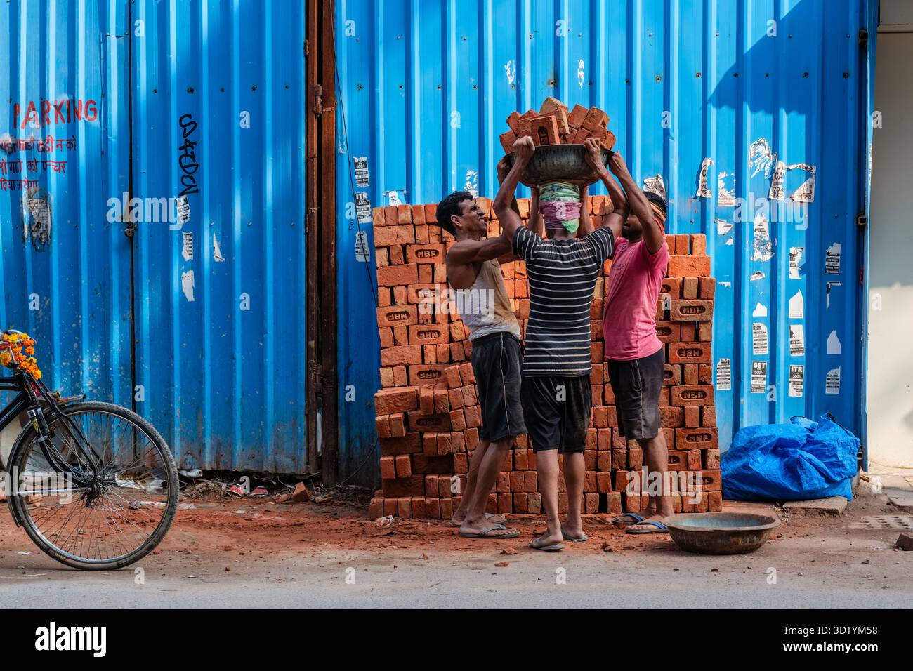 Trois ouvriers empilant des briques orange contre une clôture en métal ondulé bleu à New Delhi, avec un vélo garé sur la gauche. Banque D'Images