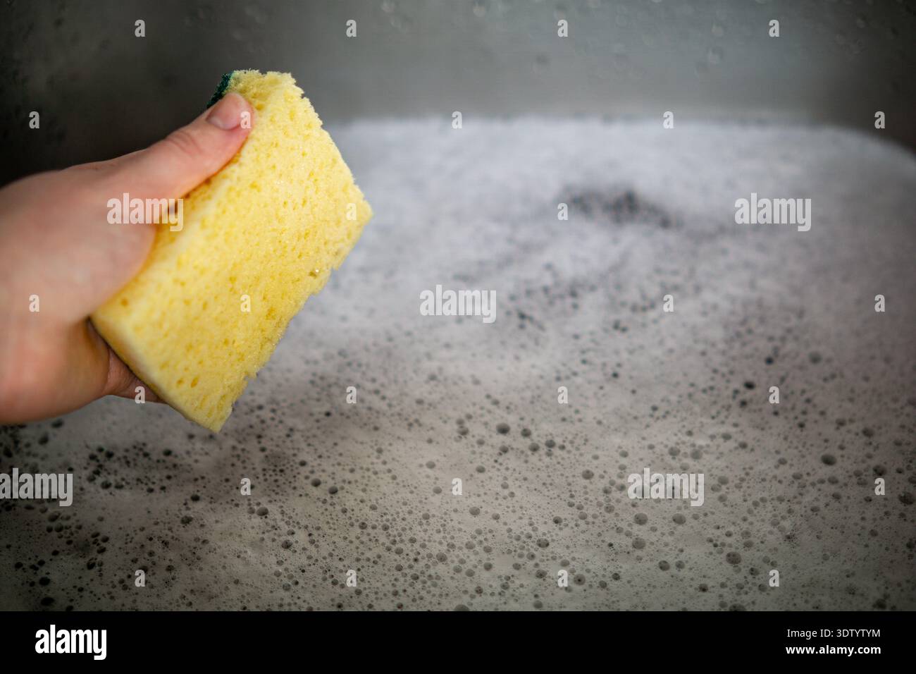 Vue rapprochée d'une main tenant une éponge de cuisine poreuse au-dessus d'un évier rempli de mousse de savon. Banque D'Images