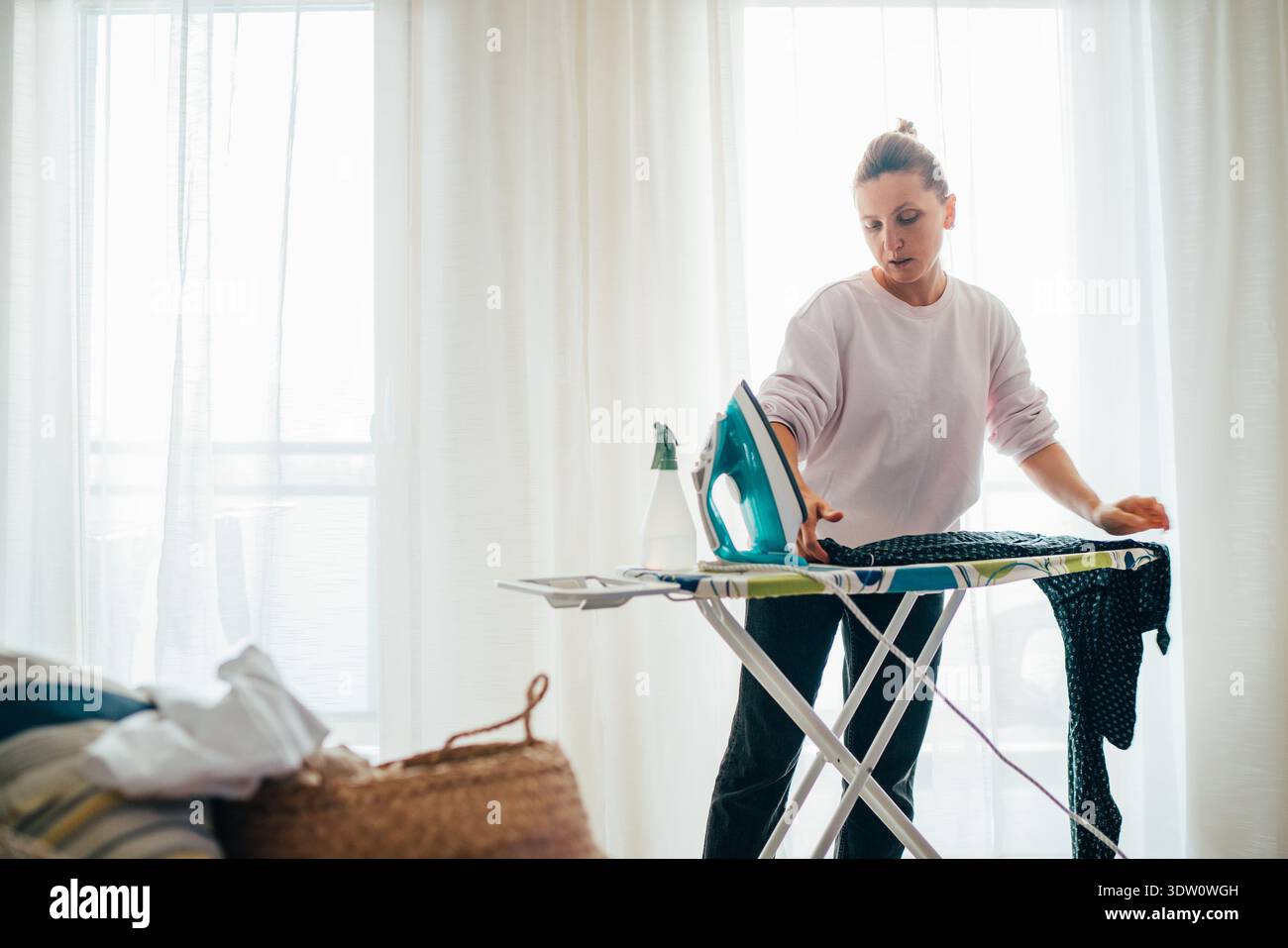 Jeune femme consciente repassant diligemment un vêtement sombre sur une planche à repasser dans une pièce lumineuse et ensoleillée, pratiquant les soins ménagers, la gestion du ménage, un Banque D'Images