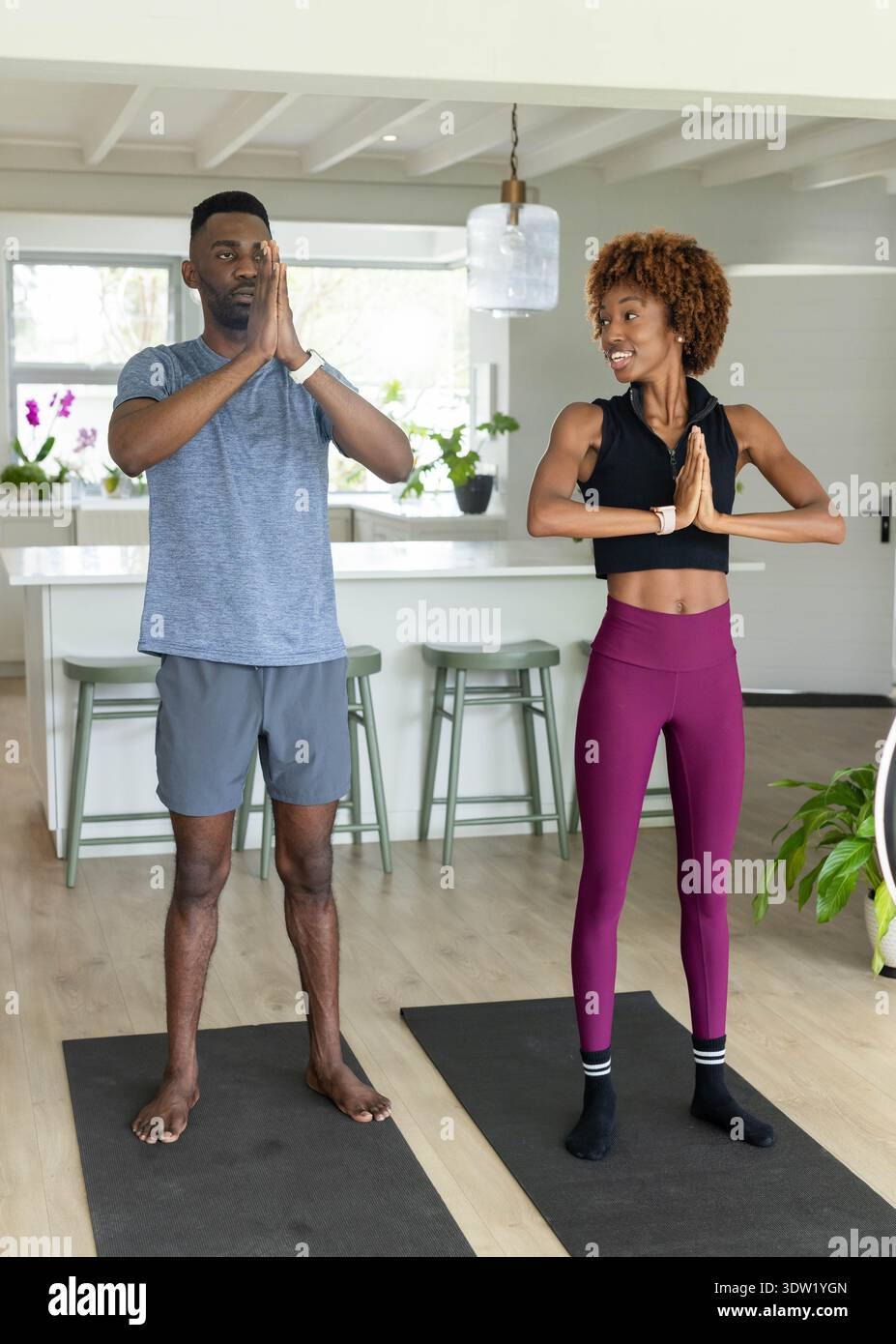 Couple diversifié pratiquant le yoga debout sur des tapis dans la cuisine, portant des vêtements de sport et des montres Banque D'Images