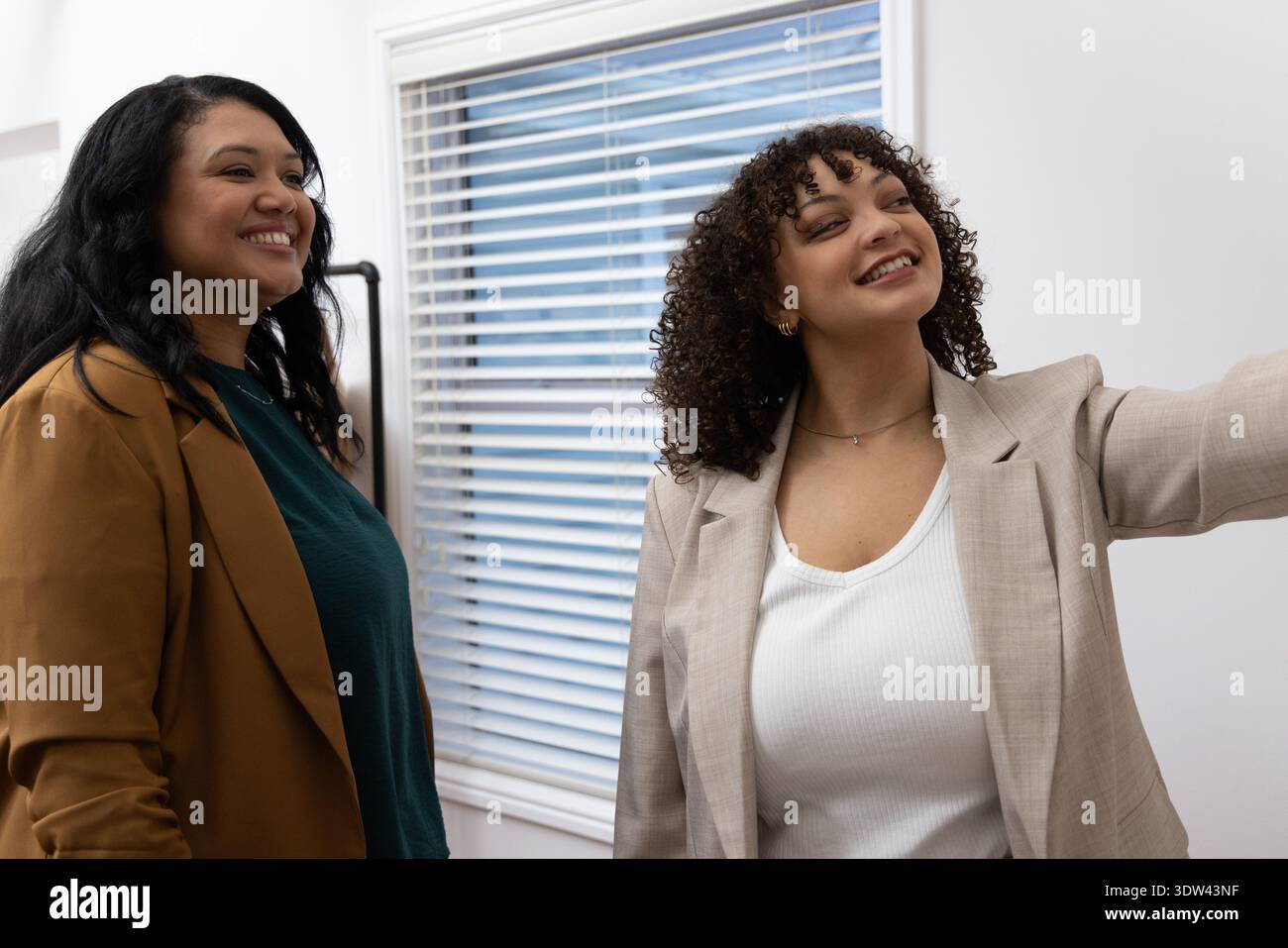 Diverses collègues féminines souriantes et debout ensemble dans le bureau avec blazers, stores, rack Banque D'Images