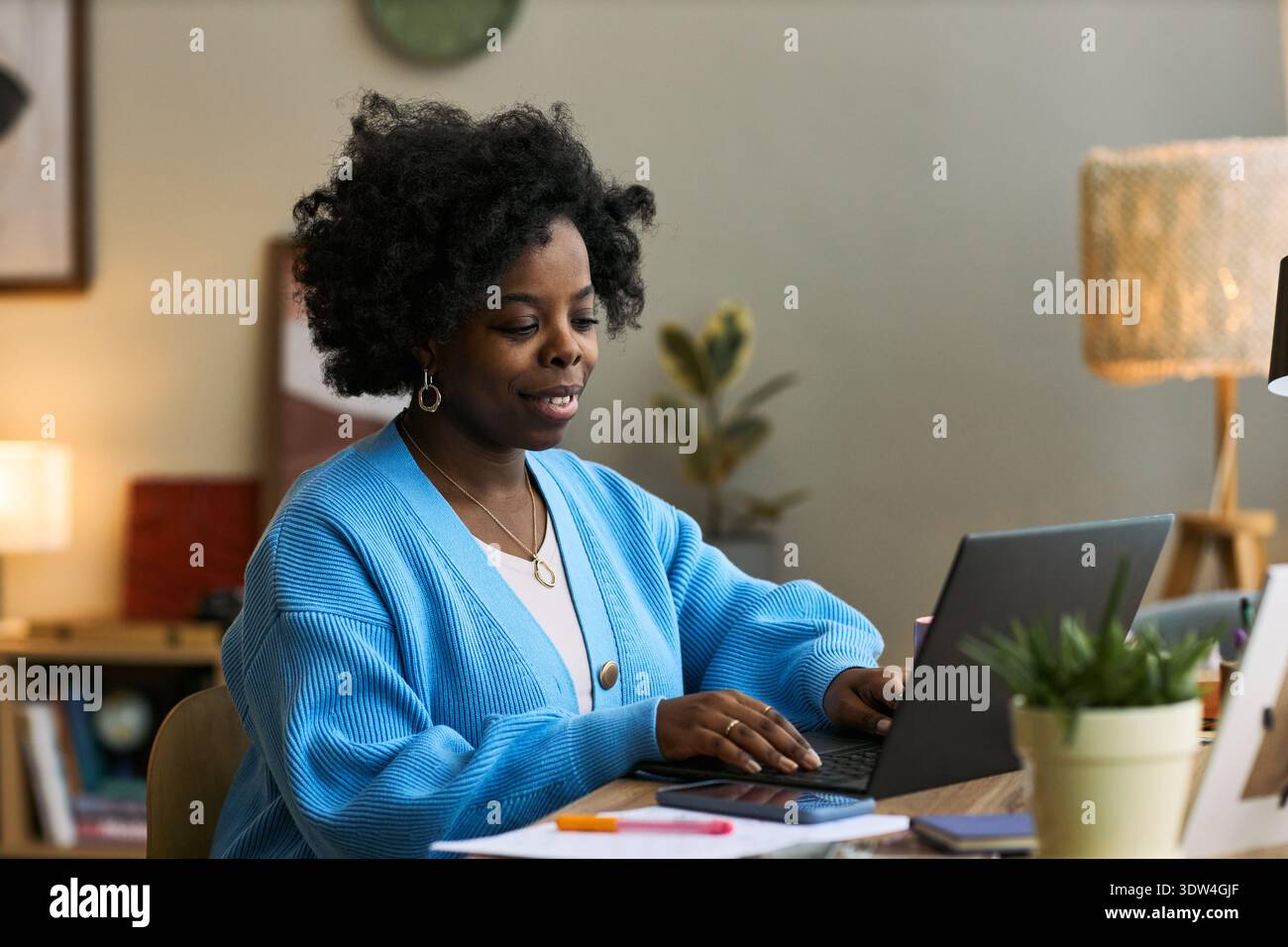 Jeune femme noire adulte travaillant sur ordinateur portable au bureau, s'engageant avec la plate-forme de santé femtech, se concentrant sur la gestion numérique de la santé, entourée de fournitures de bureau à domicile et de plantes Banque D'Images