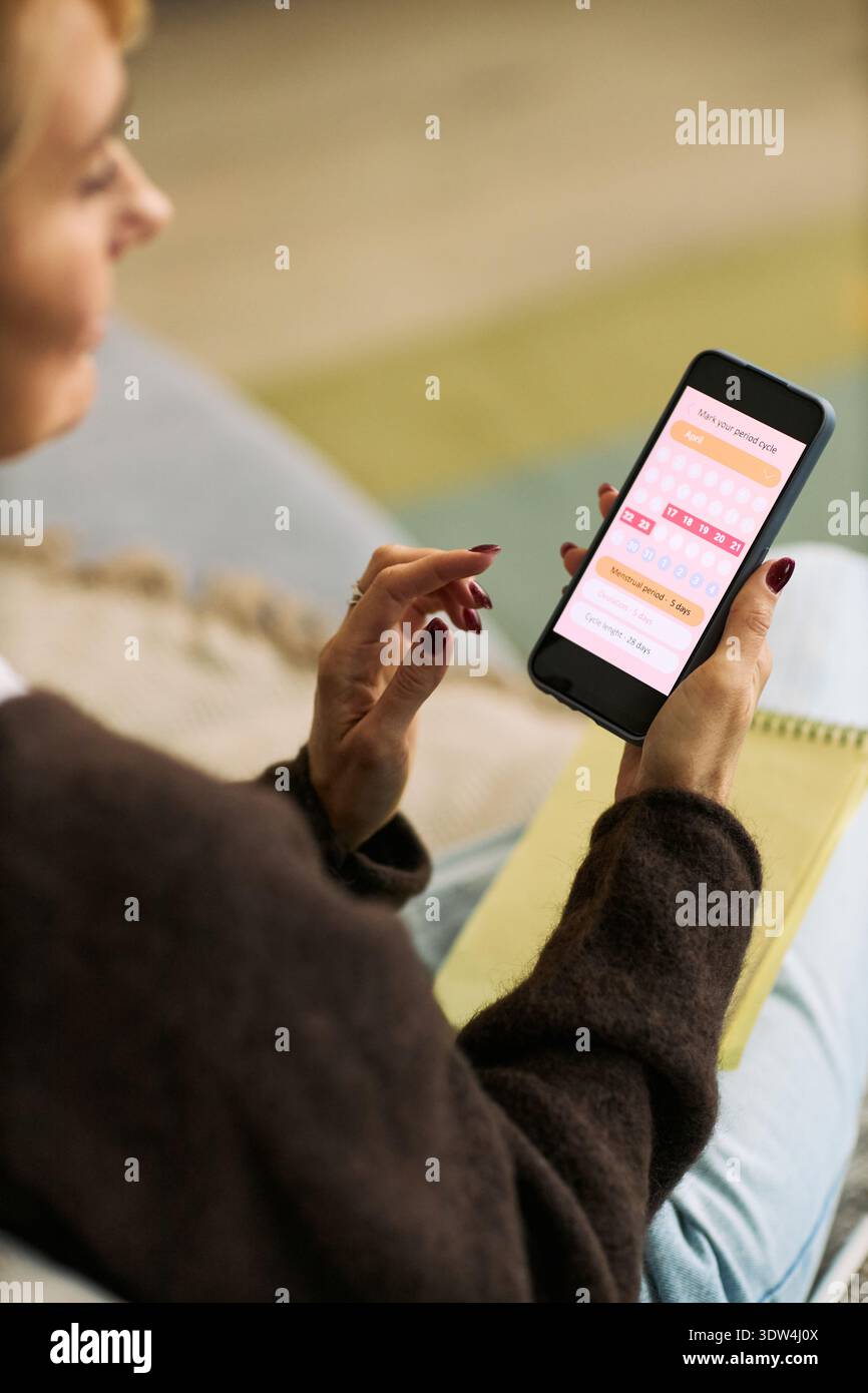 Femme caucasienne d'âge moyen utilisant l'application de suivi de la santé smartphone tout en étant assise à l'intérieur, tenant le téléphone dans une main et l'ordinateur portable dans l'autre, en se concentrant sur la technologie femtech pour le bien-être Banque D'Images