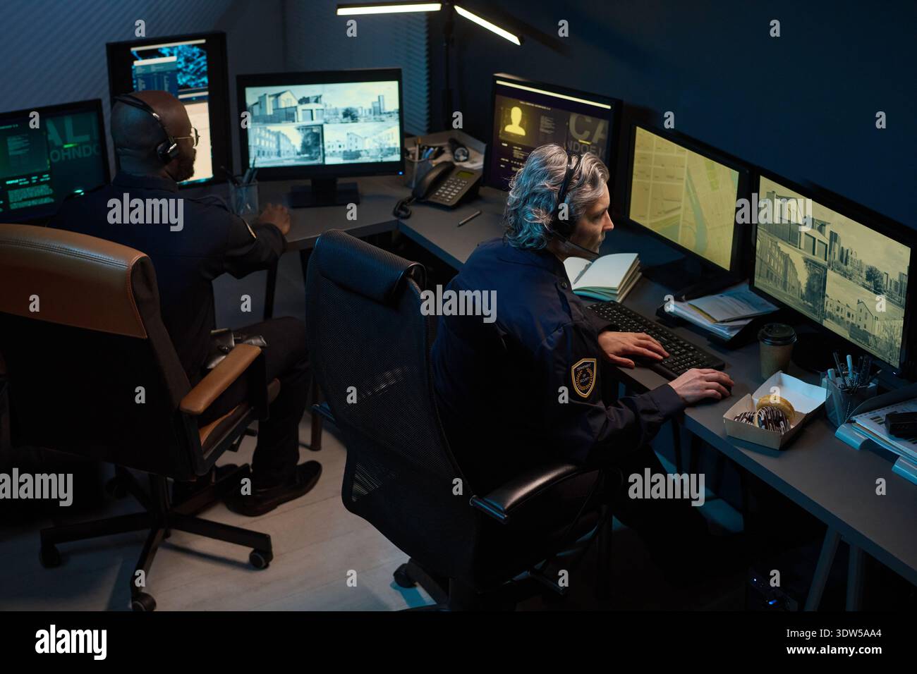 Femme caucasienne d'âge moyen et homme noir surveillant plusieurs écrans de surveillance dans la salle de contrôle de sécurité, tous deux concentrés sur l'observation des flux vidéo en direct et l'analyse des informations sur les moniteurs Banque D'Images