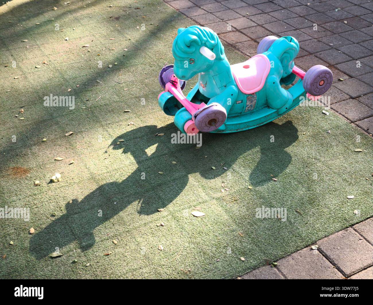 Un jouet de cheval à bascule coloré pour enfants est assis seul sur une surface de jeu extérieure, projetant une longue ombre dans la lumière du soleil du matin Banque D'Images
