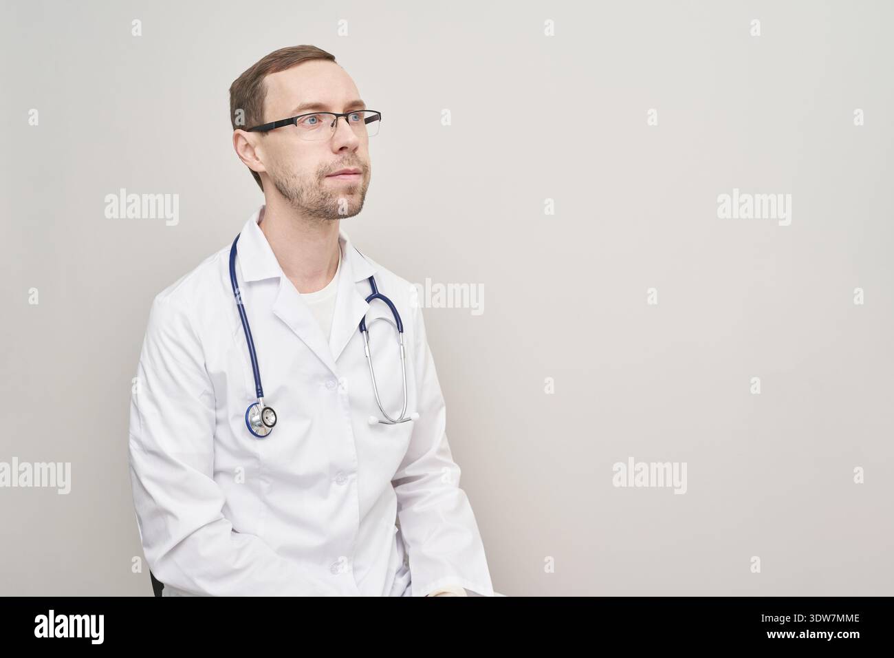 Médecin masculin caucasien en manteau blanc avec stéthoscope regardant avec confiance sur le côté dans le cadre médical professionnel. Banque D'Images