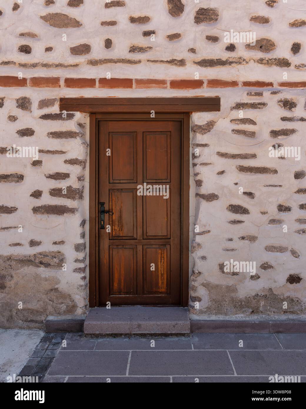 Porte en bois rustique à panneaux marron avec poignée noire dans un vieux mur en pierre plâtrée blanche avec pierres apparentes. Passerelle pavée devant. Façade de bâtiment traditionnelle Banque D'Images
