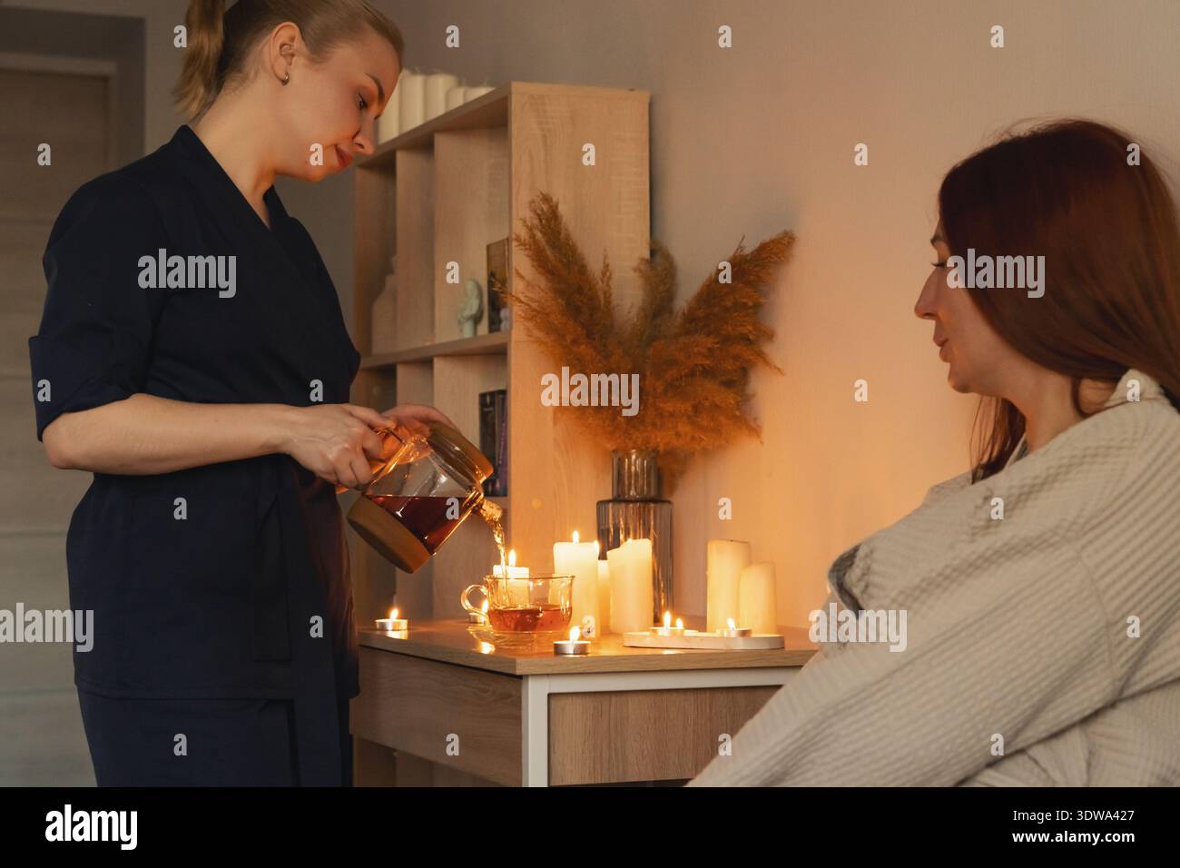 Femme thérapeute versant du thé chaud d'une théière en verre dans une tasse, offrant une cérémonie du thé apaisante dans un environnement de spa avec des bougies Banque D'Images