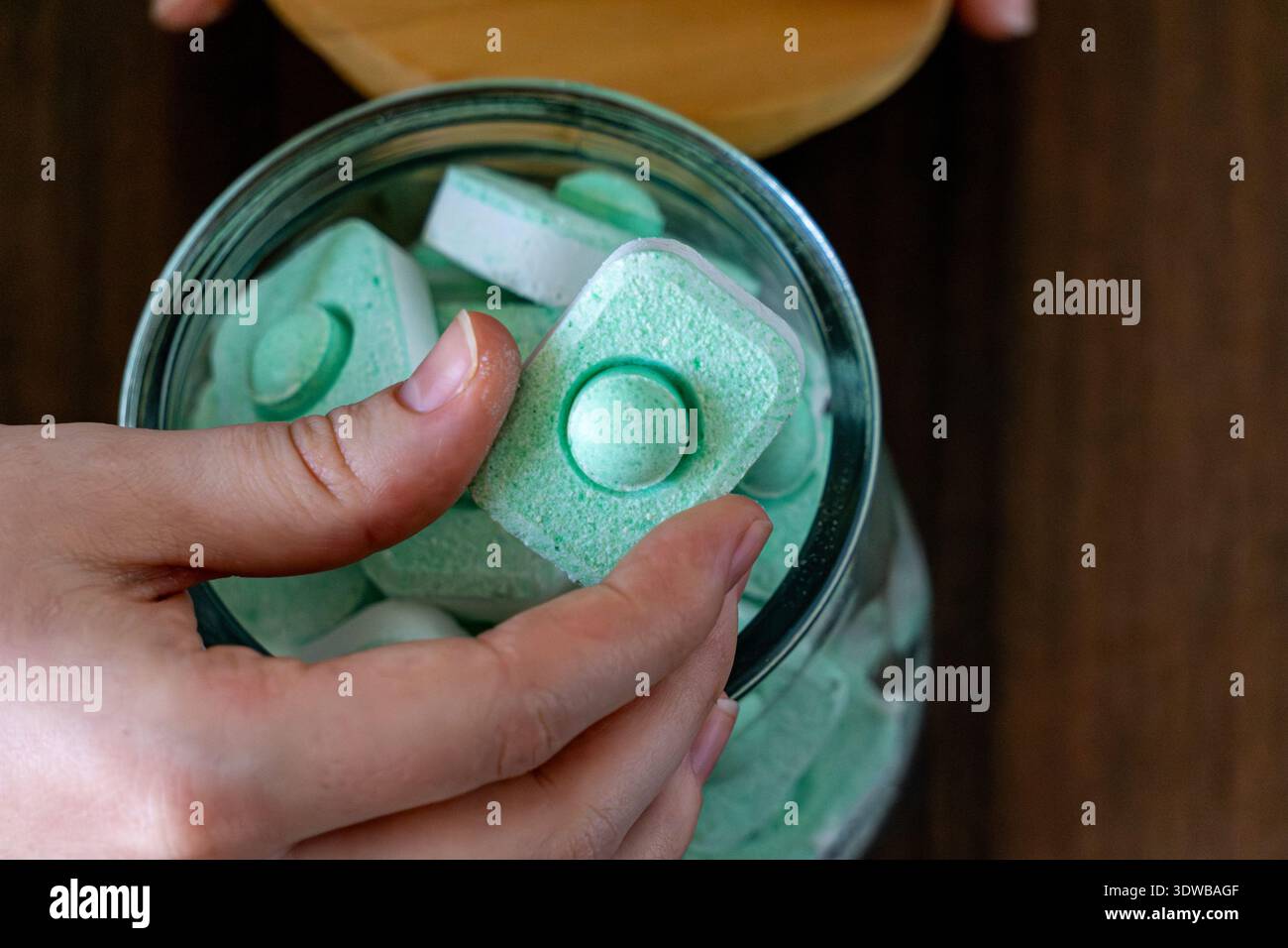 Vue de dessus gros plan d'une main tenant une pastille verte pour lave-vaisselle au-dessus d'un bocal en verre rempli de pastilles de nettoyage. Banque D'Images