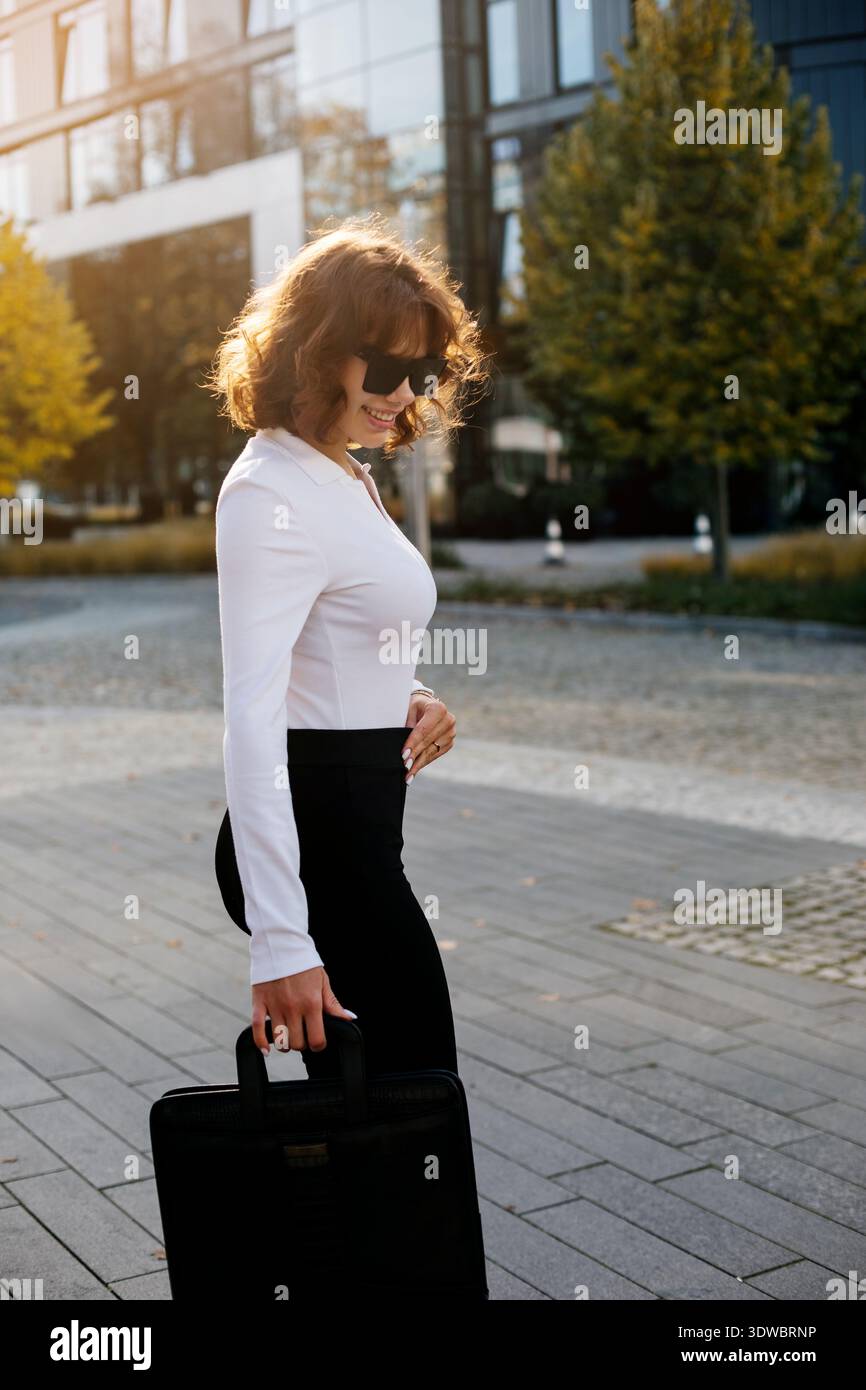 Femme d'affaires élégante dans une tenue formelle posant avec une mallette noire devant l'architecture de bureau moderne. Banque D'Images