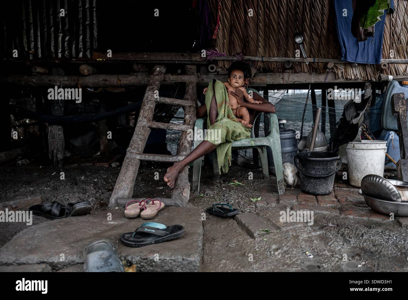 Satkhira, Bangladesh - 13 juillet 2025 : vue d'un enfant tenu dans les bras sur une chaise sous une habitation sur pilotis, une scène reflétant une vie simple au bord de l'eau Banque D'Images