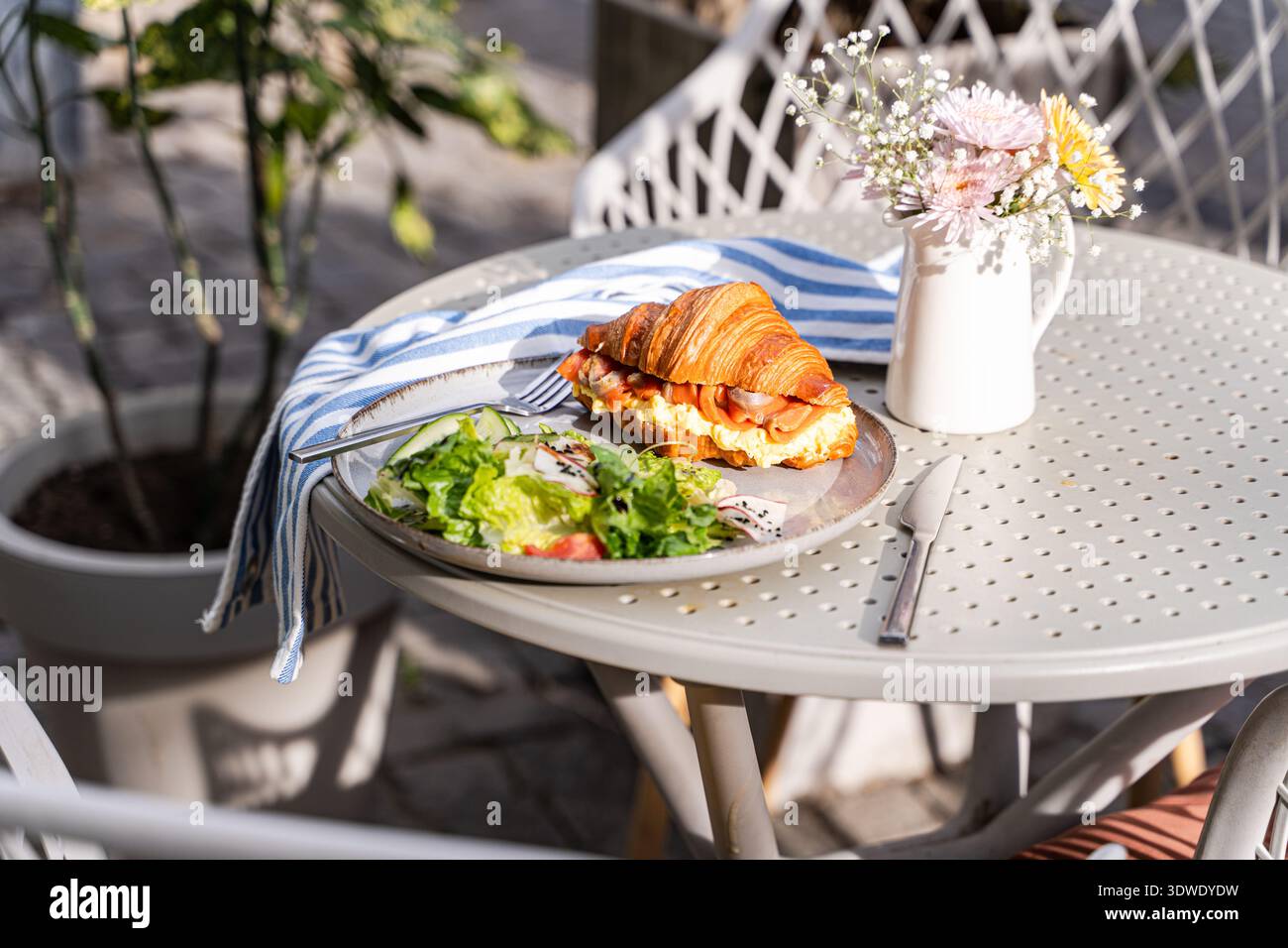 Un délicieux croissant de saumon accompagné d'une salade fraîche est servi sur une table de café ensoleillée. La scène capture un cadre parfait pour une appétizine saine Banque D'Images