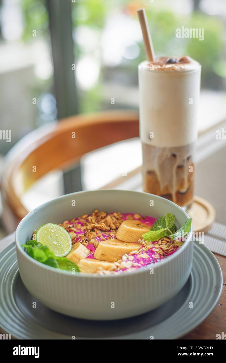 Un bol à smoothie vibrant avec des tranches de bananes fraîches, du citron vert, du granola et des feuilles de menthe est servi avec une boisson crémeuse. Parfait pour un petit déjeuner sain. Banque D'Images