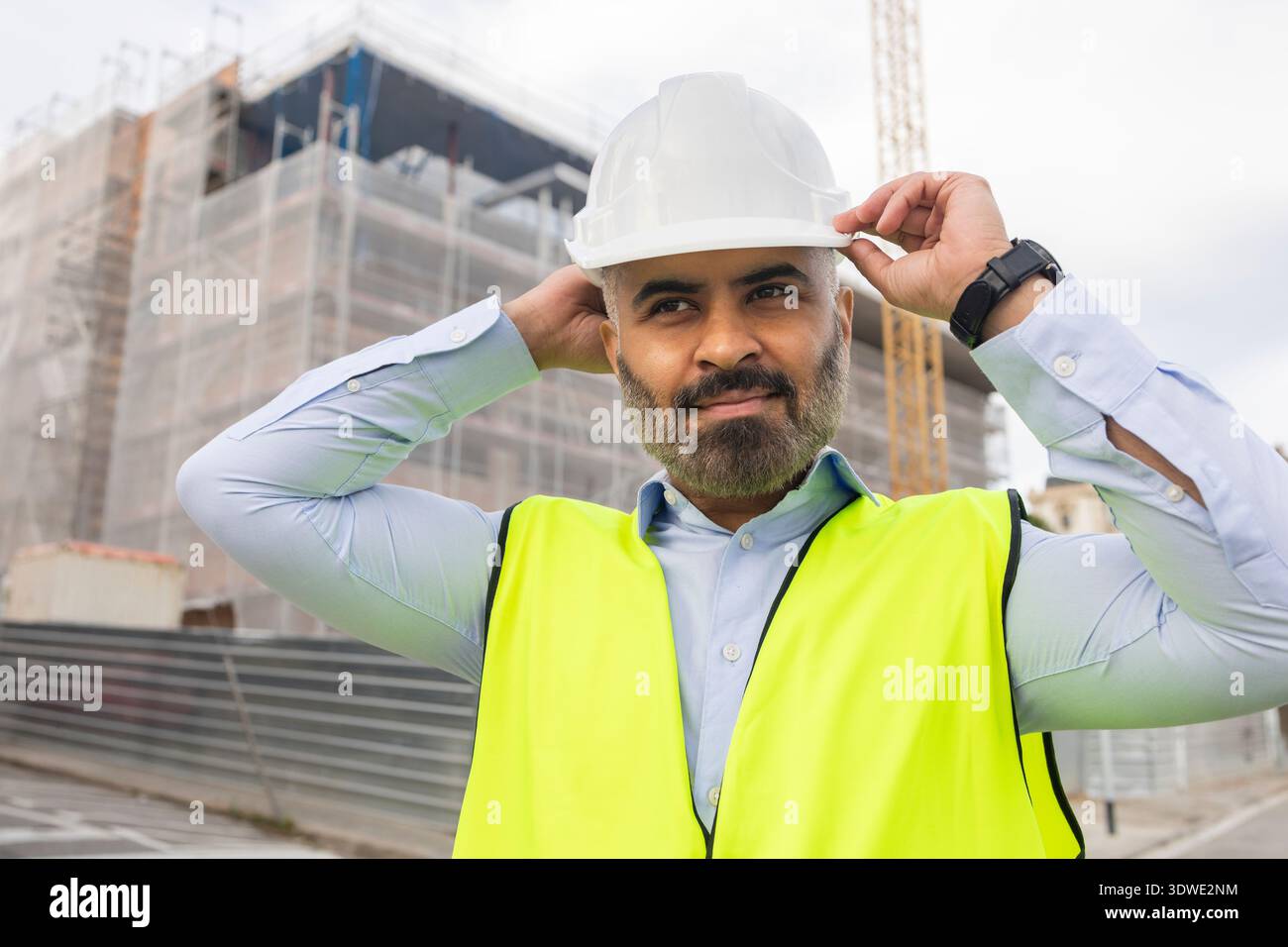 Un ingénieur portant un casque blanc et un gilet néon ajuste son équipement sur un chantier de construction occupé. Derrière lui, un projet de construction est activement en cours Banque D'Images