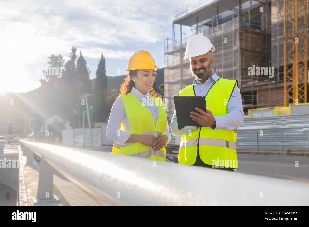 Deux ingénieurs en construction portant des gilets de sécurité et des casques examinent les plans du site sur une tablette. Ils se tiennent près d'un chantier de construction urbain, mettant en valeur le travail d'équipe Banque D'Images
