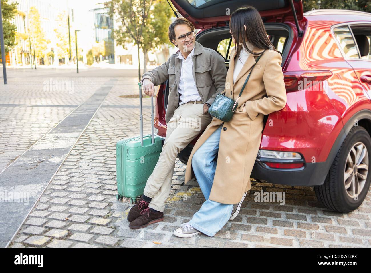 Un couple se prépare pour un Road trip, appuyé contre leur voiture rouge avec un coffre ouvert. Le cadre de la rue et leur style moderne mettent en évidence un spontané Banque D'Images